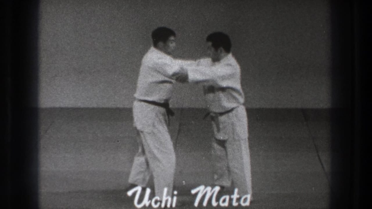 The two adult practitioners in white gi begin the Uchi Mata movement, gripping near the arms and shoulders in the indoor training space.