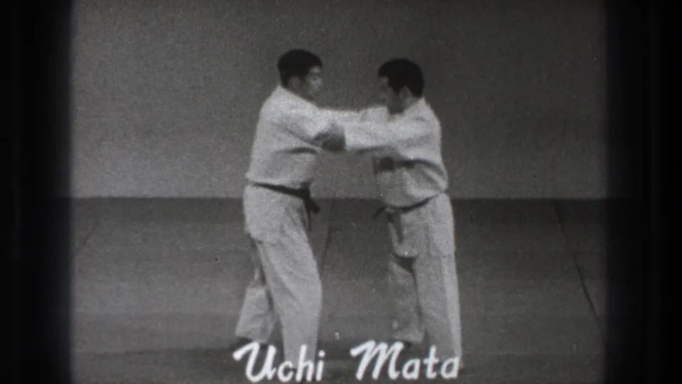 The two adult practitioners in white gi begin the Uchi Mata movement, gripping near the arms and shoulders in the indoor training space.