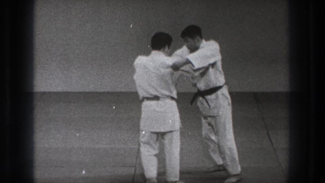 The martial arts pair continue the standing Uchi Mata demonstration, with one man facing away and the other braced in front of him on the mat.