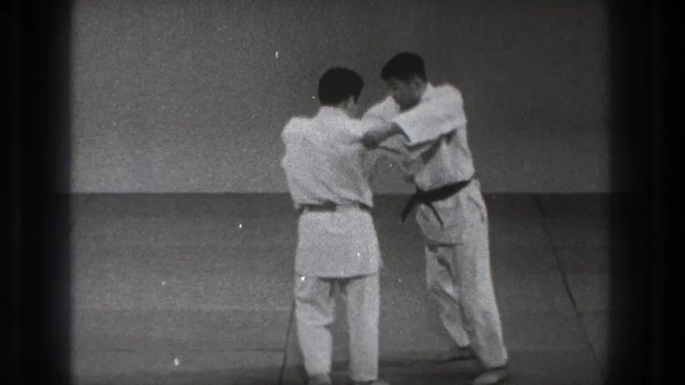 The martial arts pair continue the standing Uchi Mata demonstration, with one man facing away and the other braced in front of him on the mat.