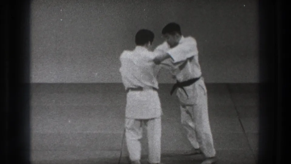 In 1971 Tokyo footage, two men in white gi stand on mats while one begins positioning for a judo throw.