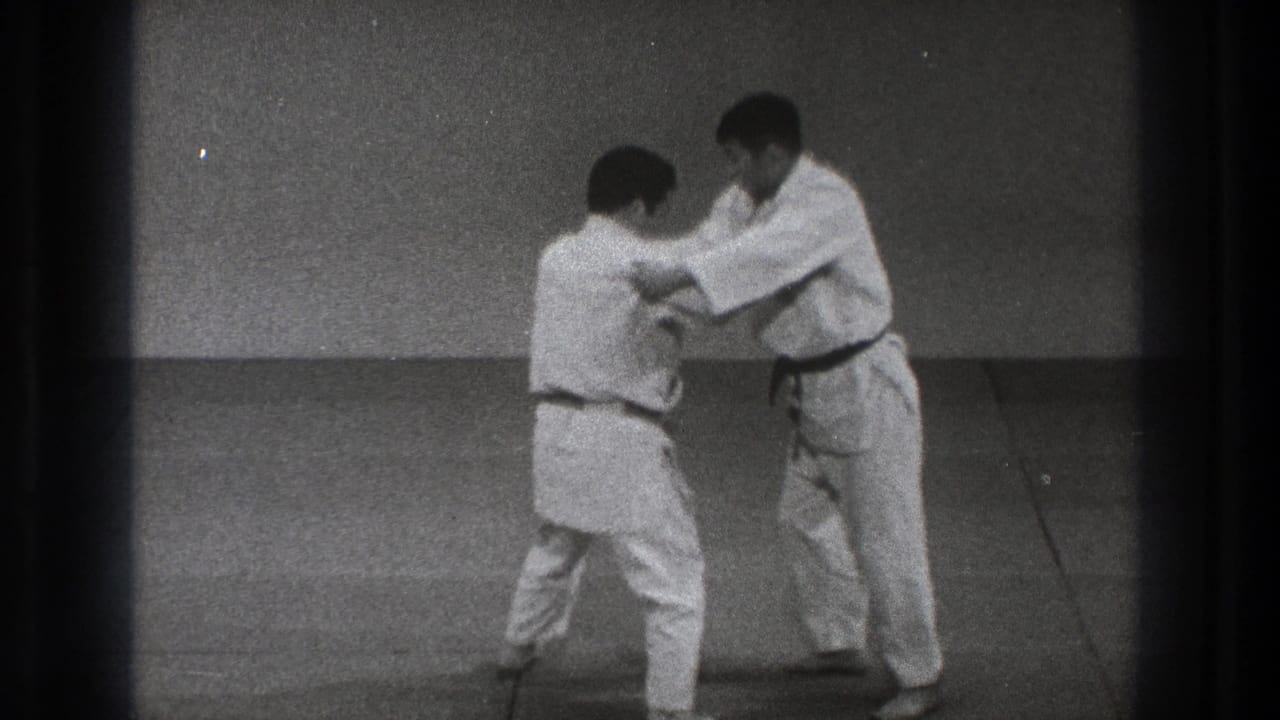 Two men in white gi continue a controlled judo practice exchange on mats in the 1971 Tokyo clip.