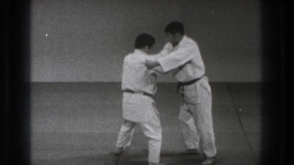 Two men in white gi continue a controlled judo practice exchange on mats in the 1971 Tokyo clip.