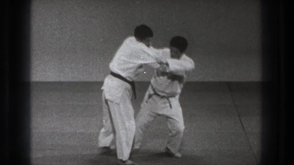 In supplied 1971 Tokyo footage, two men in white gi stand on a simple mat as one begins controlling the other's arm and jacket.