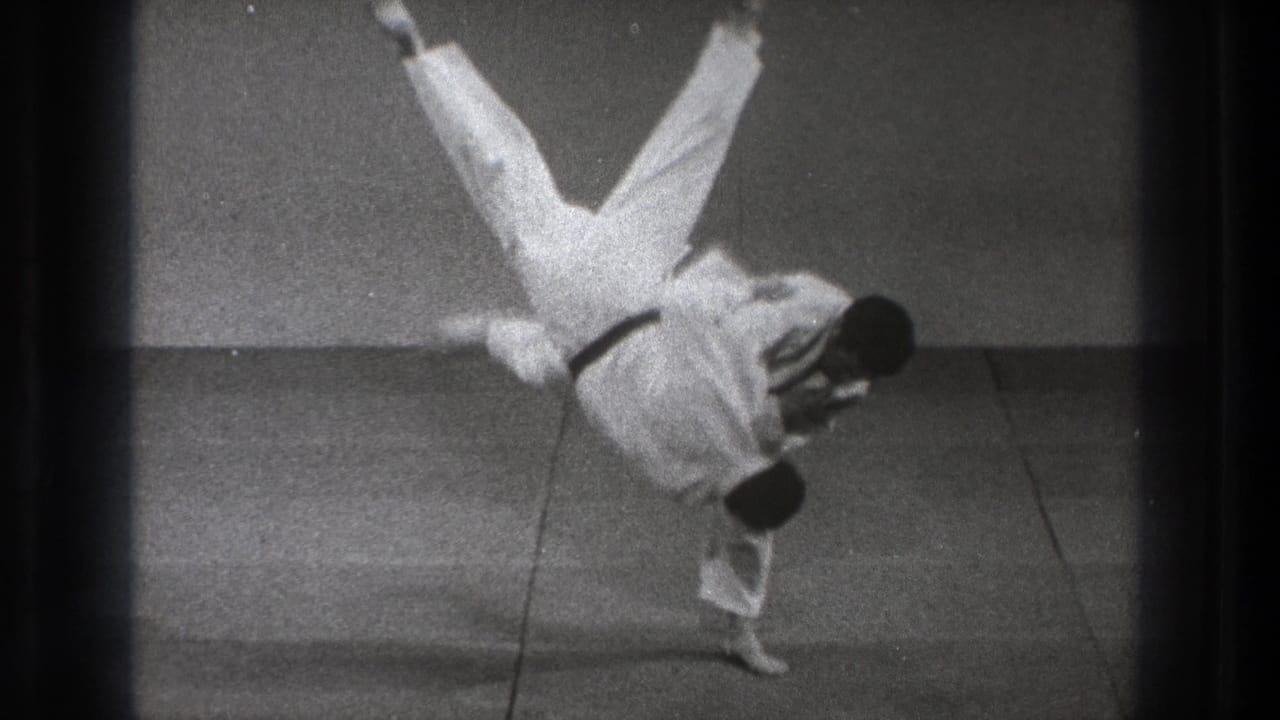 In the final moment, the thrown practitioner flips upside down over the black-belt man's shoulder on the matted floor.