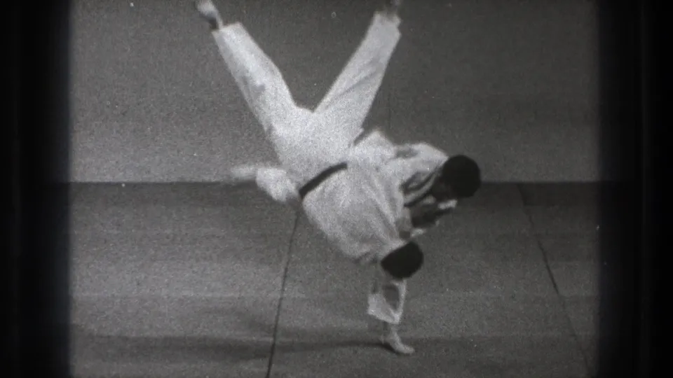 In the final moment, the thrown practitioner flips upside down over the black-belt man's shoulder on the matted floor.