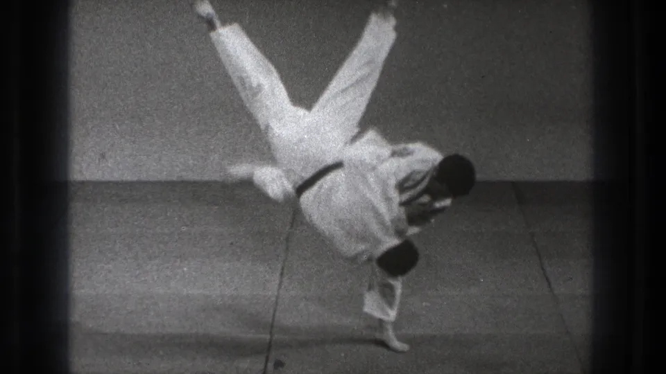 In 1971 Tokyo footage, one man in a white gi is lifted or flipped while another supports him during a martial arts demonstration.