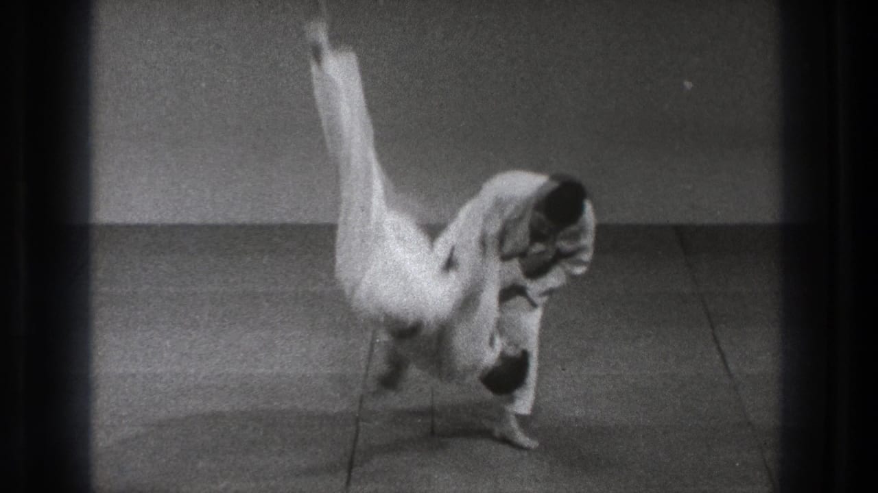 A black-and-white 8mm frame shows a white-uniformed practitioner rotating through a high flip during a Tokyo karate instruction sequence.