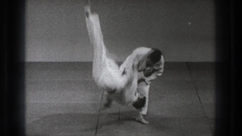 A black-and-white 8mm frame shows a white-uniformed practitioner rotating through a high flip during a Tokyo karate instruction sequence.