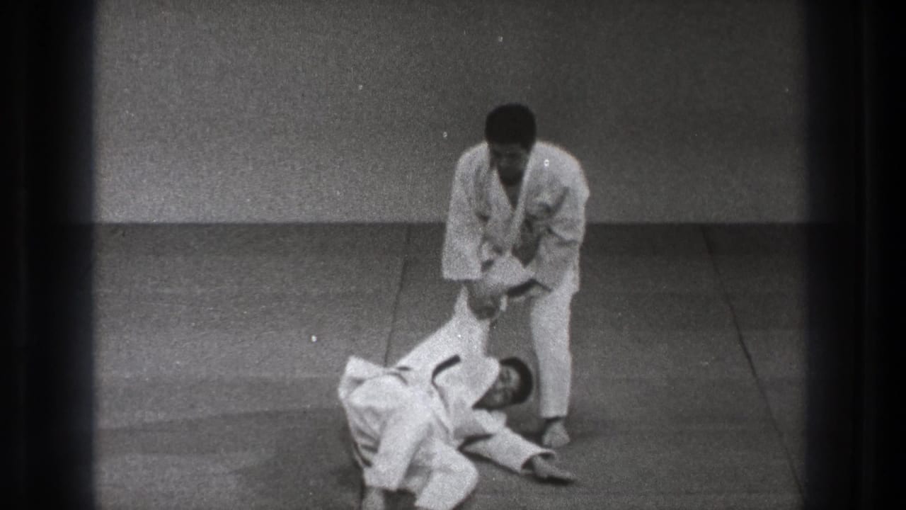 A Tokyo martial arts instruction frame shows one man standing over a partner on the mat while maintaining an arm grip after the throw.