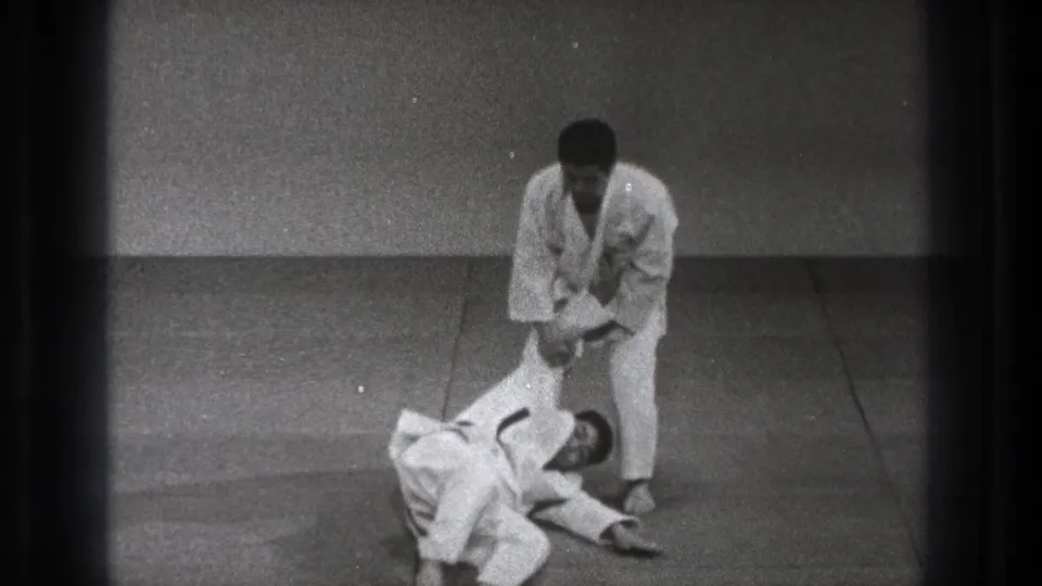 A Tokyo martial arts instruction frame shows one man standing over a partner on the mat while maintaining an arm grip after the throw.