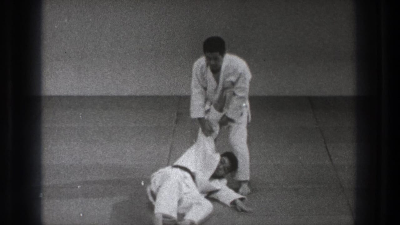 Two men in white gi practice a throw on an indoor mat.