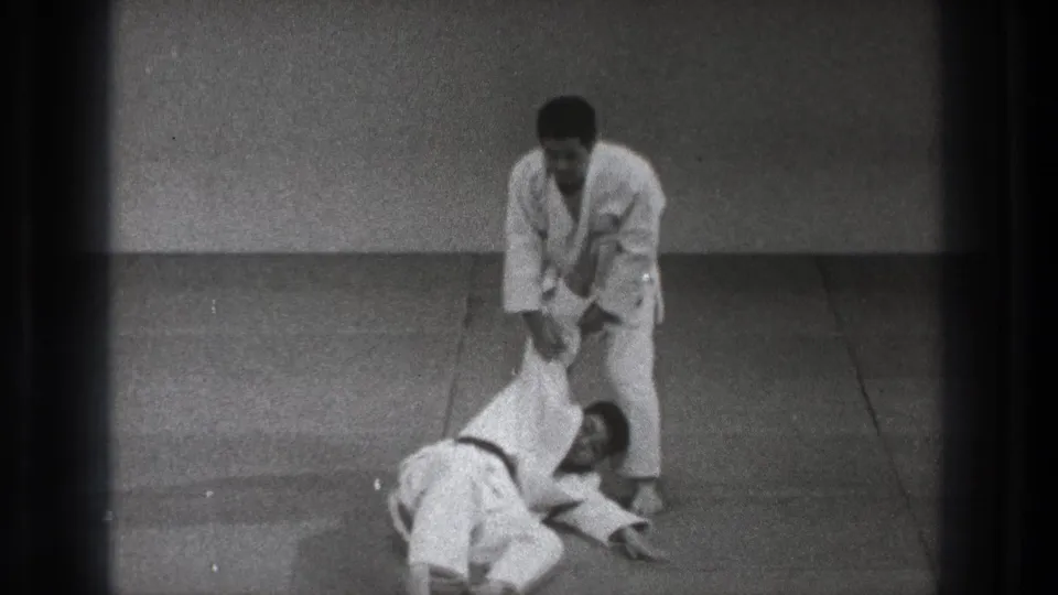 In 1971 Tokyo footage, one adult in a white gi controls another practitioner during a martial arts throw on a plain indoor mat.