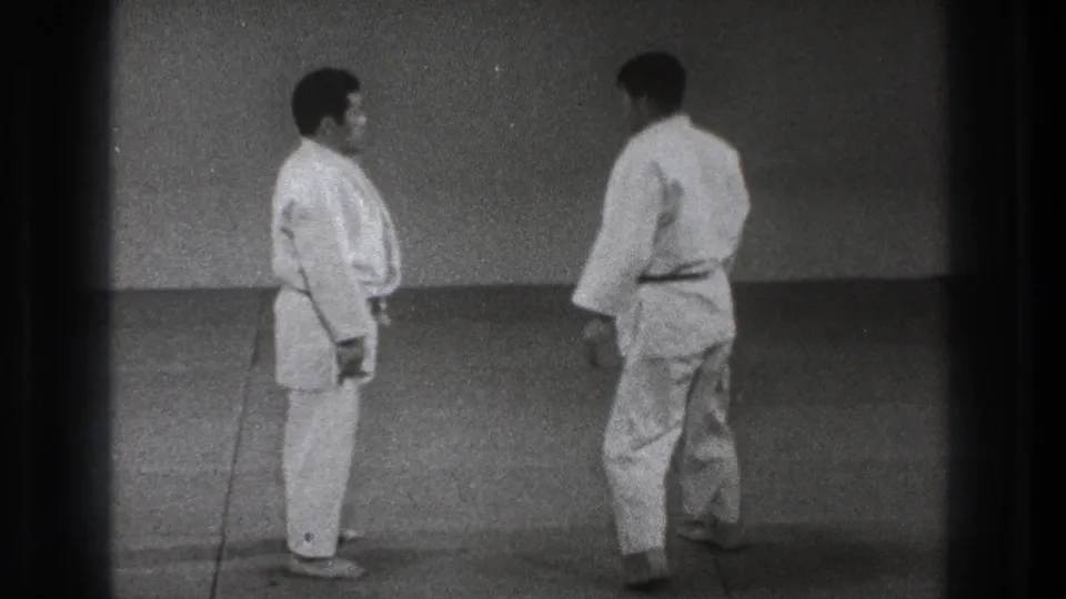 In the supported 1971 Tokyo clip, two adult men in white judo gis stand in a simple dojo-like mat space before practice.