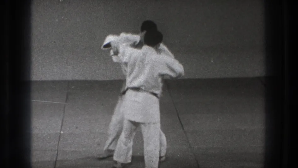 The practitioners move into close contact during a controlled judo maneuver, partly turned away from the camera.
