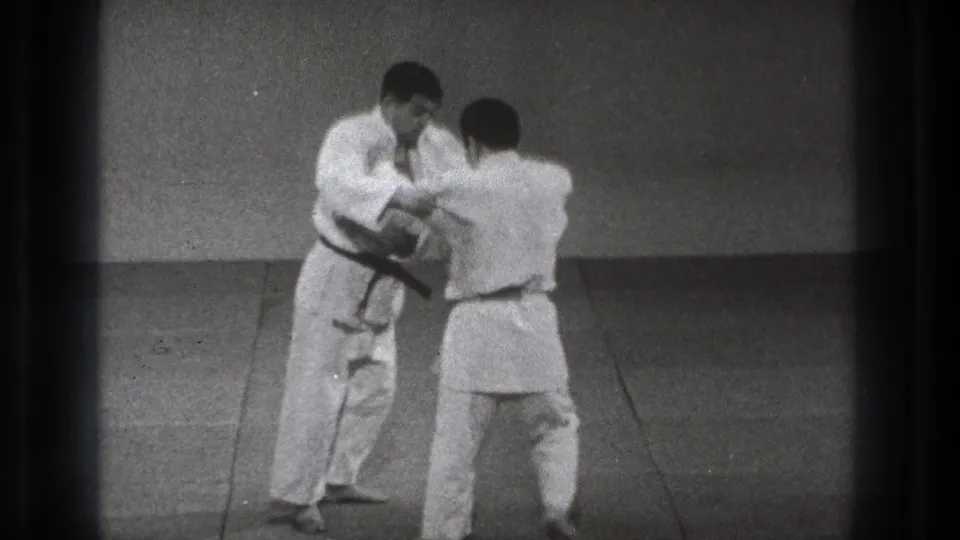 In 1971 Tokyo home-movie footage, two adult men in white judo gis begin a grappling exchange on a plain dojo mat.