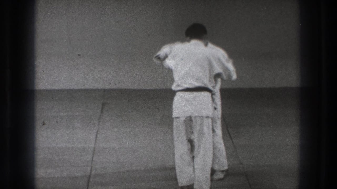 Two men in judo uniforms are seen from behind and partly obscured as they continue a throw setup on the dojo mat.