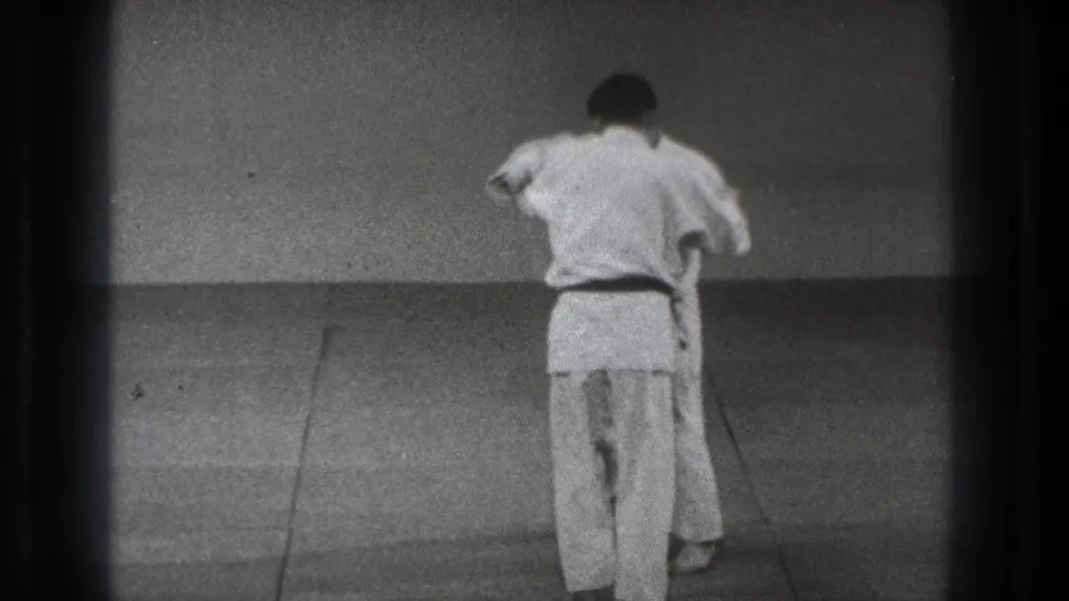 Two men in judo uniforms are seen from behind and partly obscured as they continue a throw setup on the dojo mat.