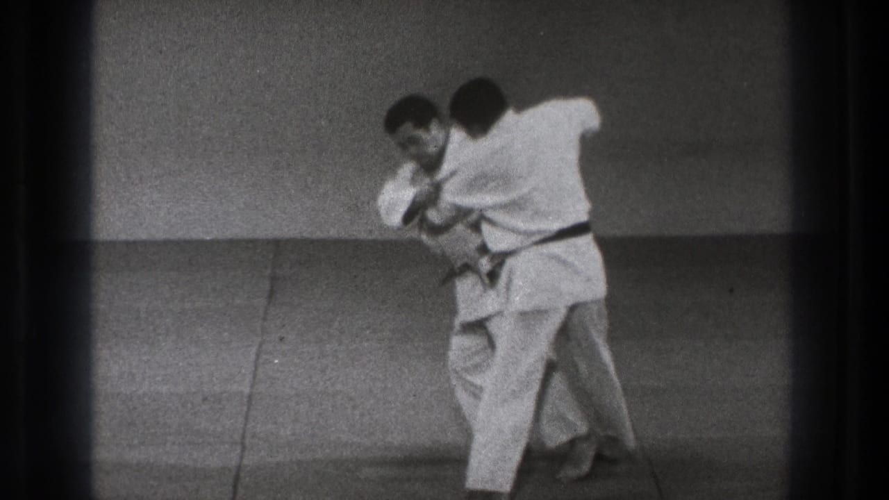 One man in a white gi turns into his partner and pulls him forward, showing a judo throw in progress on the mat.