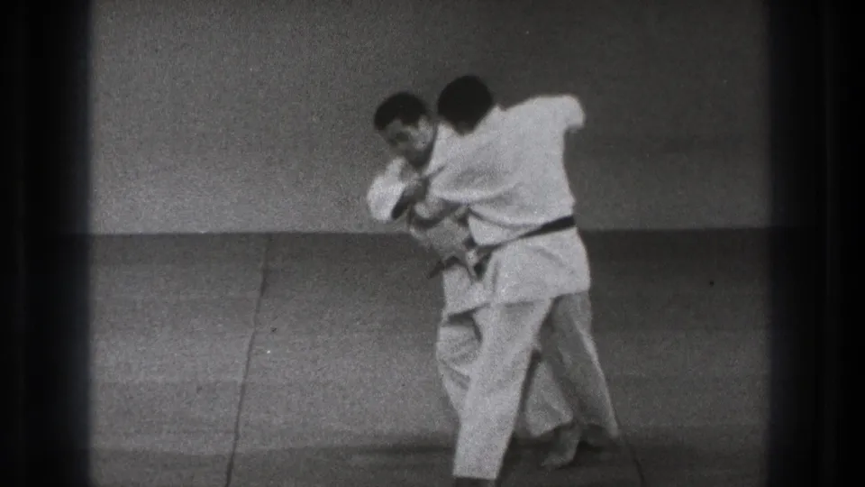 One man in a white gi turns into his partner and pulls him forward, showing a judo throw in progress on the mat.