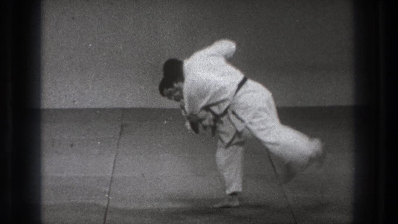 A man in a white gi executes a judo throw, lifting his partner across his hip or shoulder in the 1971 Tokyo dojo scene.