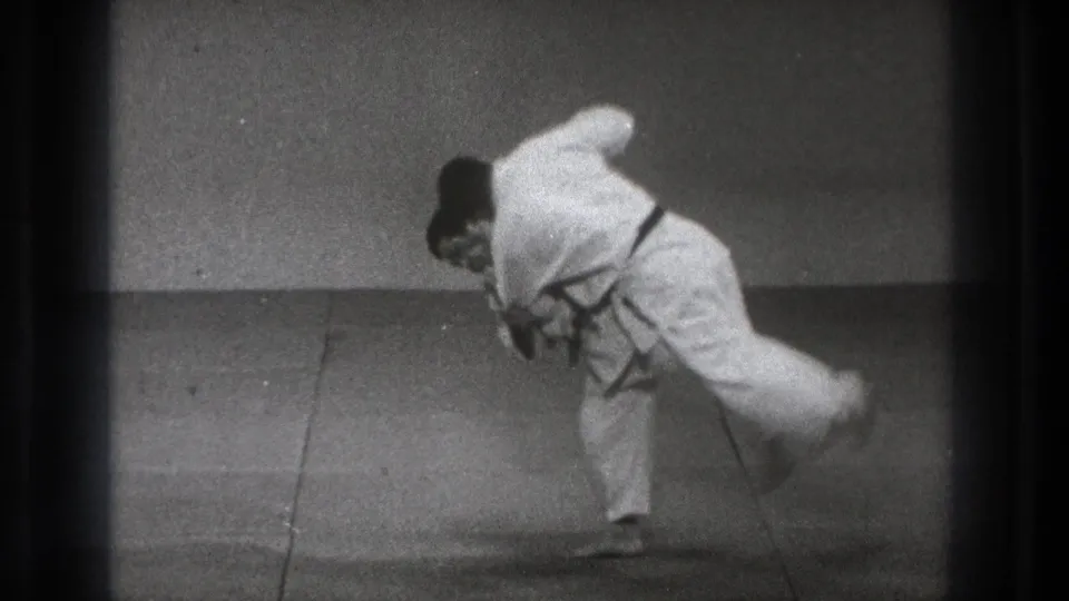 A man in a white gi executes a judo throw, lifting his partner across his hip or shoulder in the 1971 Tokyo dojo scene.