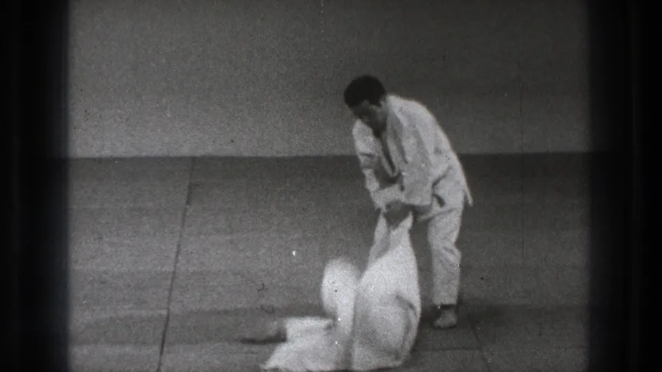 After the throw, one practitioner stands and holds the other by the gi as the opponent lies on the dojo mat.
