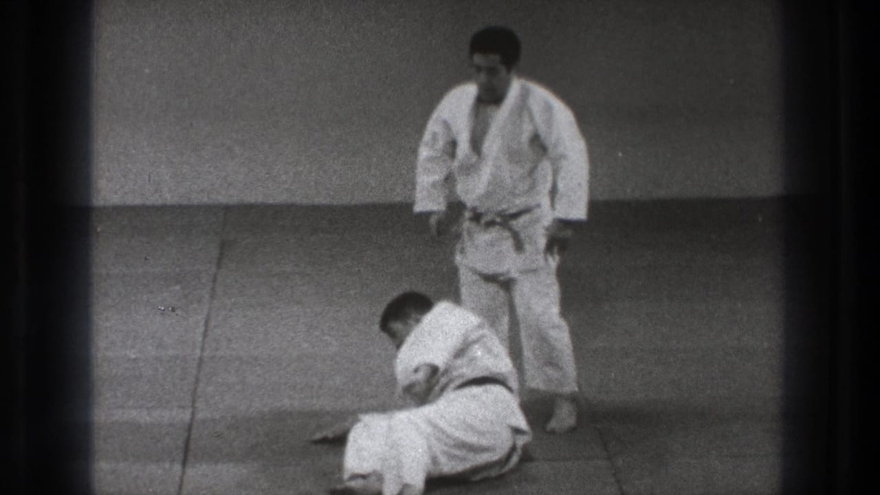 Two men in judo uniforms on a tatami mat, with one seated after a throw.