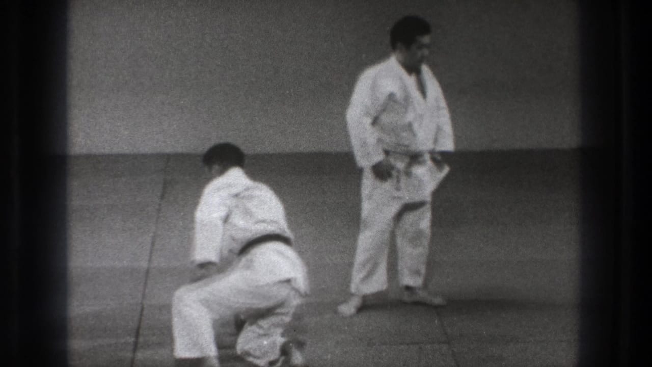 A kneeling judo practitioner begins to rise on the mat as another man in a white gi stands nearby in the Tokyo training footage.