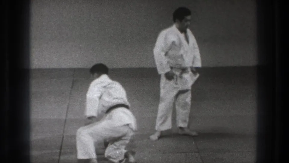 A kneeling judo practitioner begins to rise on the mat as another man in a white gi stands nearby in the Tokyo training footage.