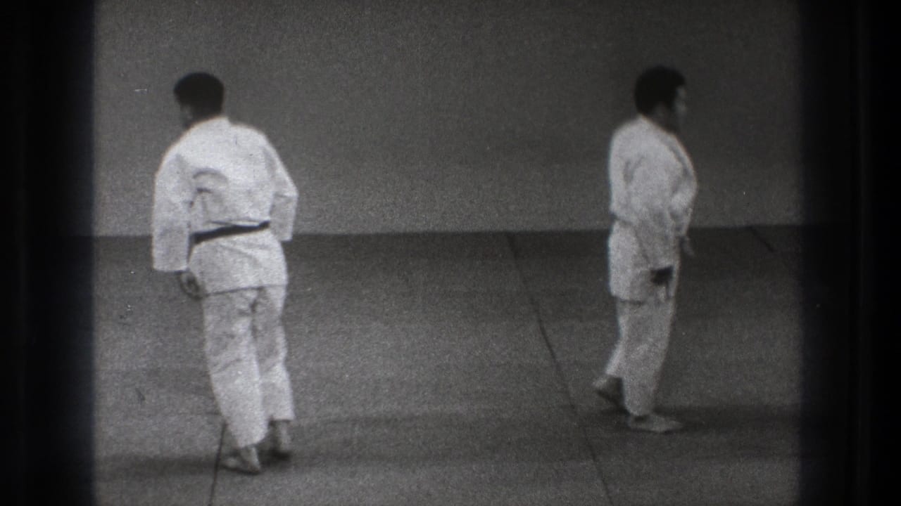 Two adult judo practitioners in white gis walk across a plain tatami practice area in the stated 1971 Tokyo home-movie clip.