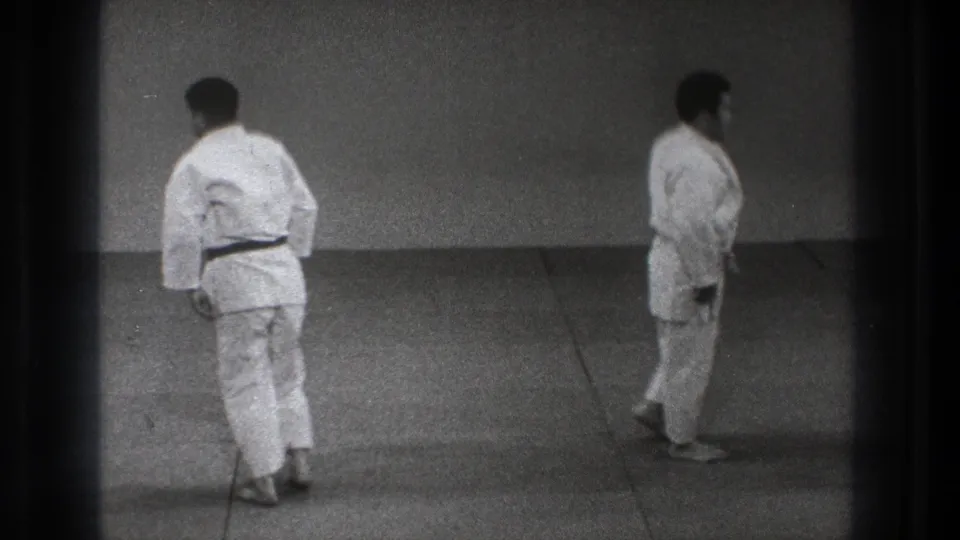 Two adult judo practitioners in white gis walk across a plain tatami practice area in the stated 1971 Tokyo home-movie clip.
