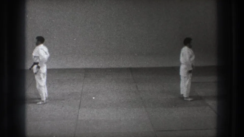 Two martial arts practitioners in white gi stand separated on the mat in Tokyo, with one dark belt visible.