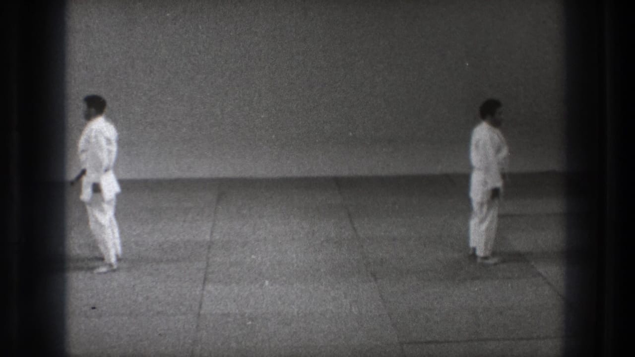 A static 8mm frame shows two men in white gi standing on a mat floor inside a plain practice space in Tokyo.
