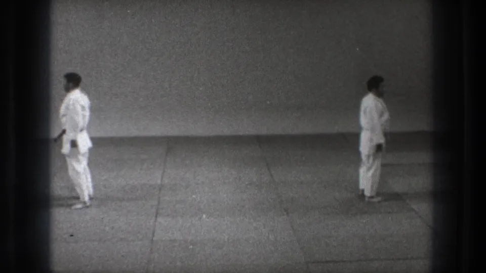 A static 8mm frame shows two men in white gi standing on a mat floor inside a plain practice space in Tokyo.