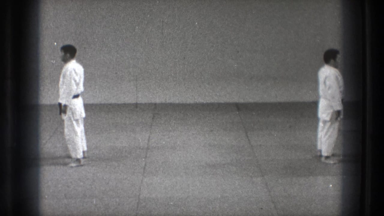 1971 Tokyo 8mm frame showing two men in white gi standing at attention on a grey dojo mat against a plain wall.
