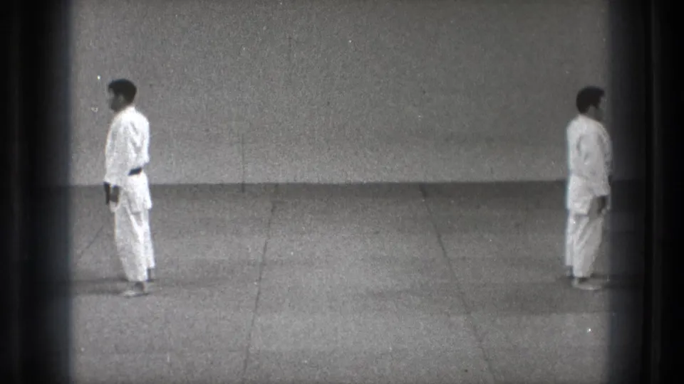 1971 Tokyo 8mm frame showing two men in white gi standing at attention on a grey dojo mat against a plain wall.
