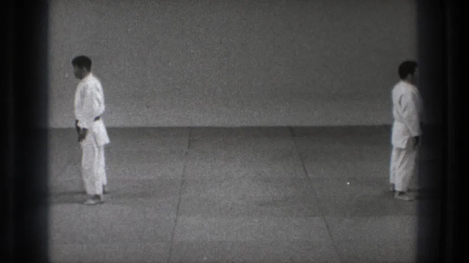 In supported 1971 Tokyo 8mm footage, two adult men in white martial arts uniforms stand separated in a plain dojo-like space.