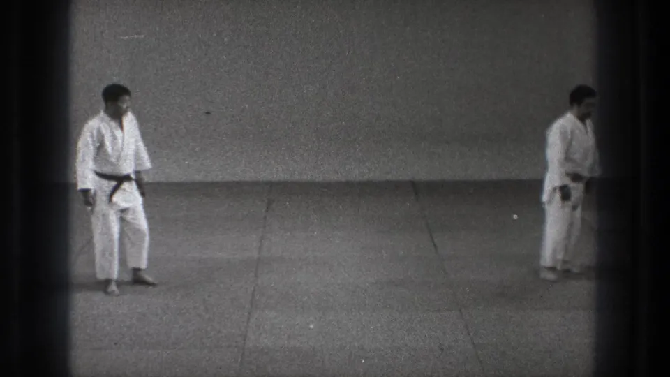 Two men in white martial arts uniforms face each other across a tiled floor in the supported Tokyo 1971 home-movie clip.