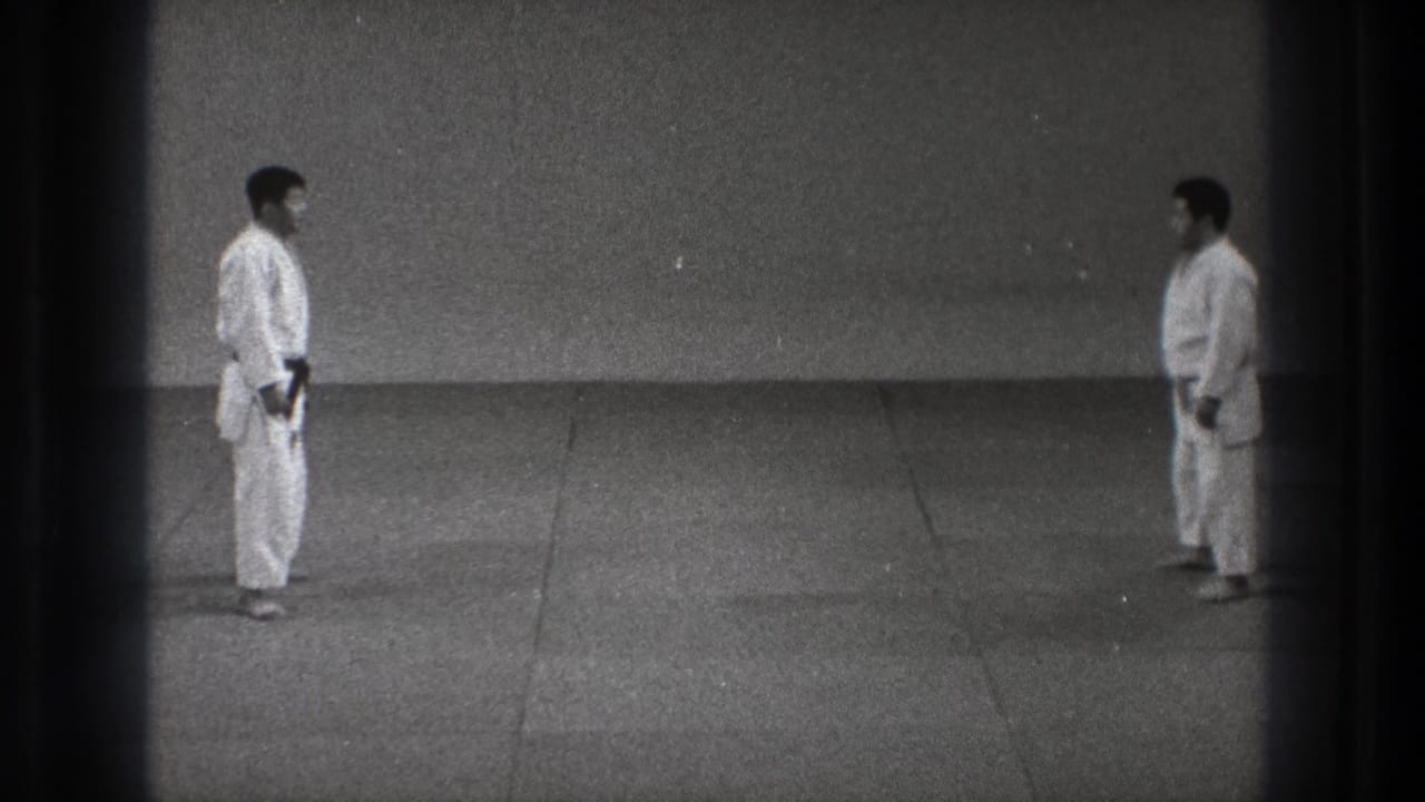 The two adult practitioners remain facing each other in the sparse Tokyo training space, one with a dark belt visible.