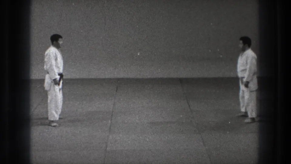 The two adult practitioners remain facing each other in the sparse Tokyo training space, one with a dark belt visible.