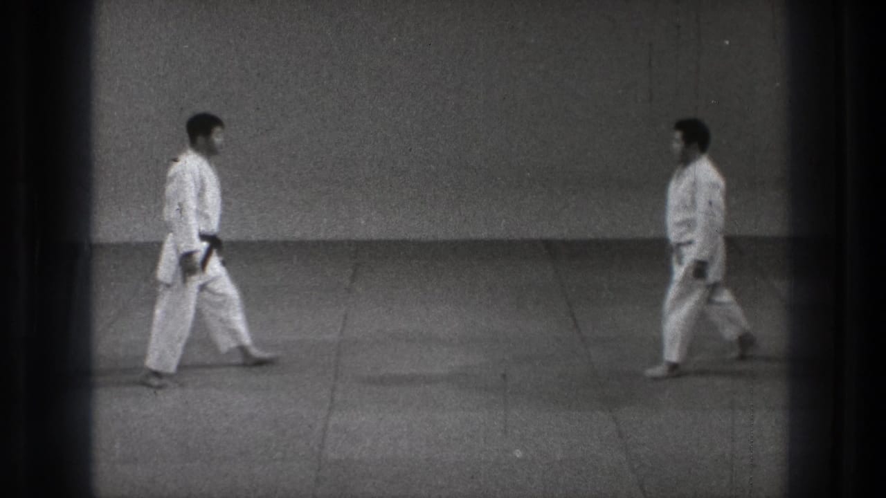 In the closing frame, two men in white martial arts uniforms approach each other across the plain indoor training floor.