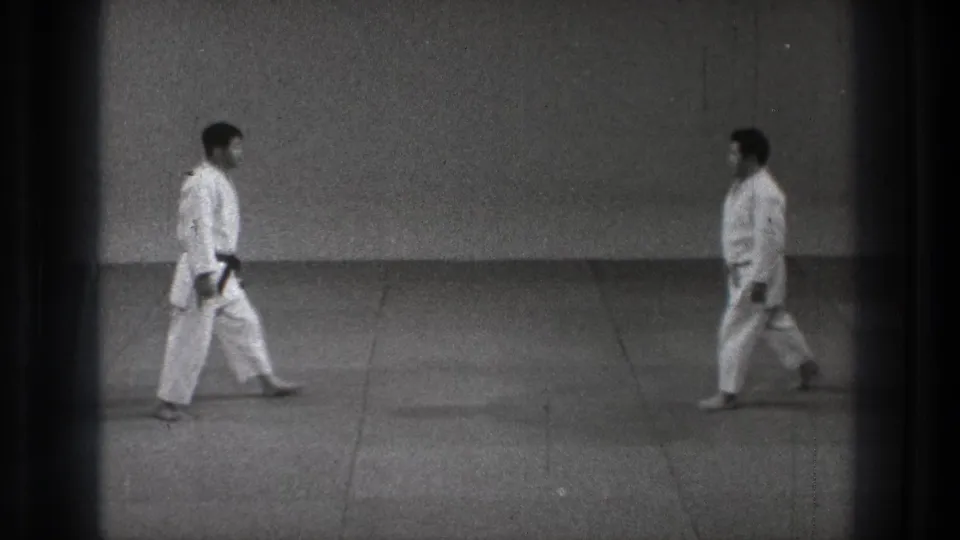 In the closing frame, two men in white martial arts uniforms approach each other across the plain indoor training floor.