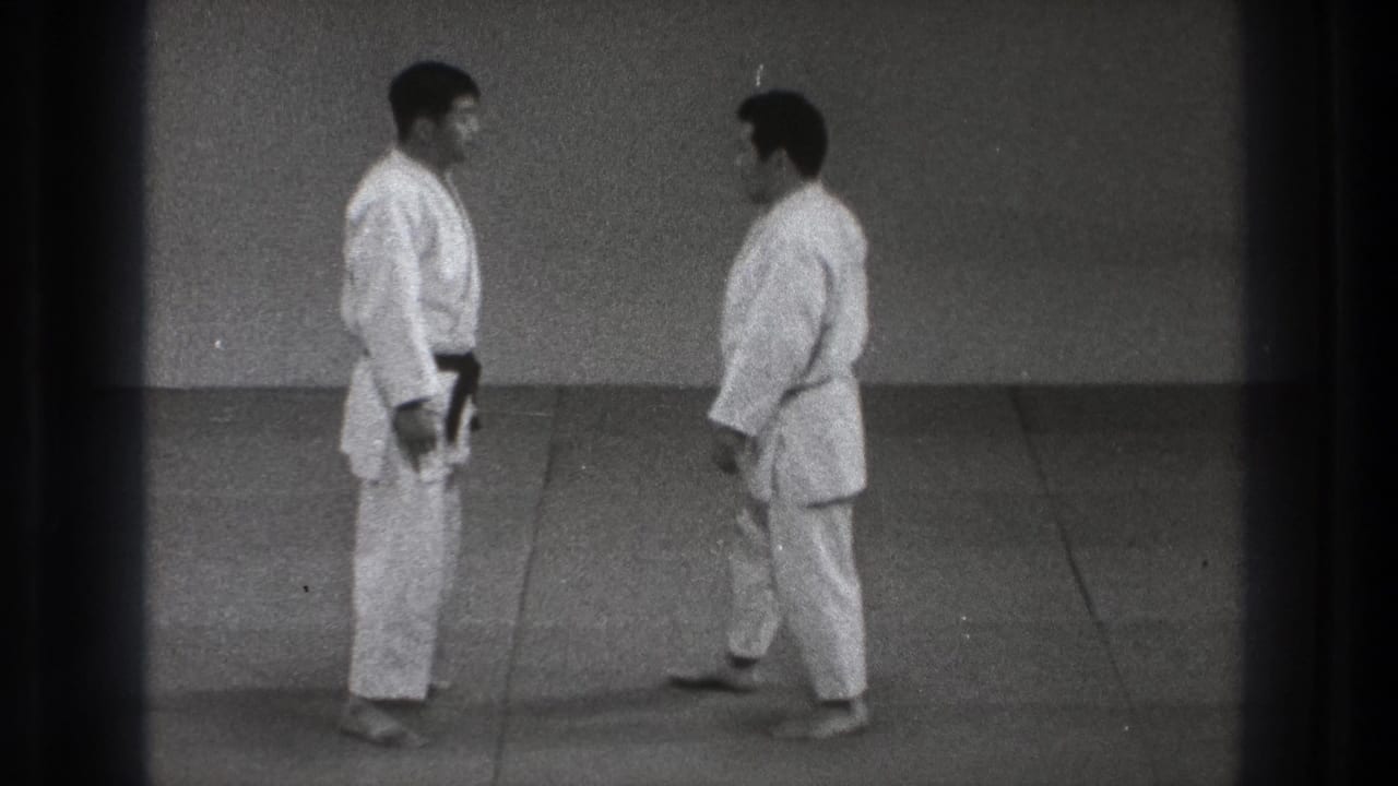 Two adult martial arts practitioners in white gi stand near each other on an indoor mat in the Tokyo 1971 clip.