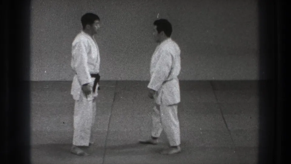 Two adult martial arts practitioners in white gi stand near each other on an indoor mat in the Tokyo 1971 clip.