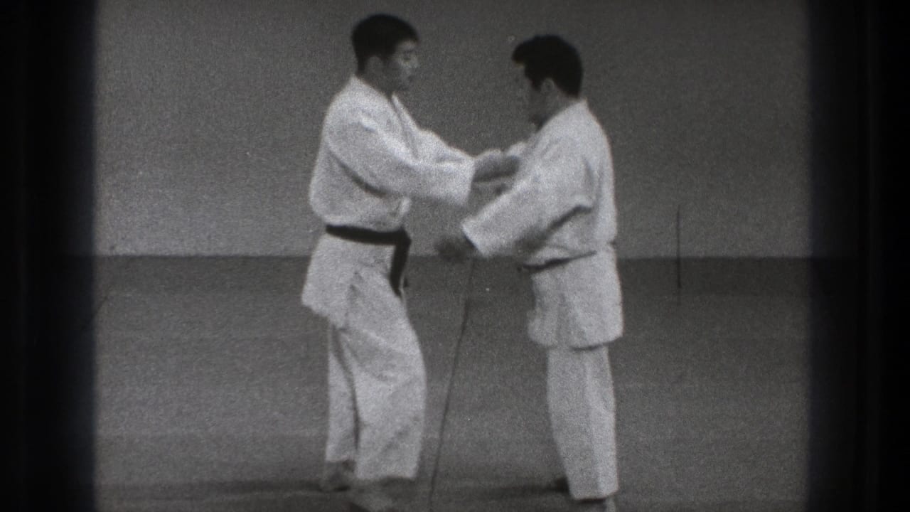 Two adult men in gi make sleeve and forearm contact during a controlled martial arts technique in the 1971 Tokyo footage.