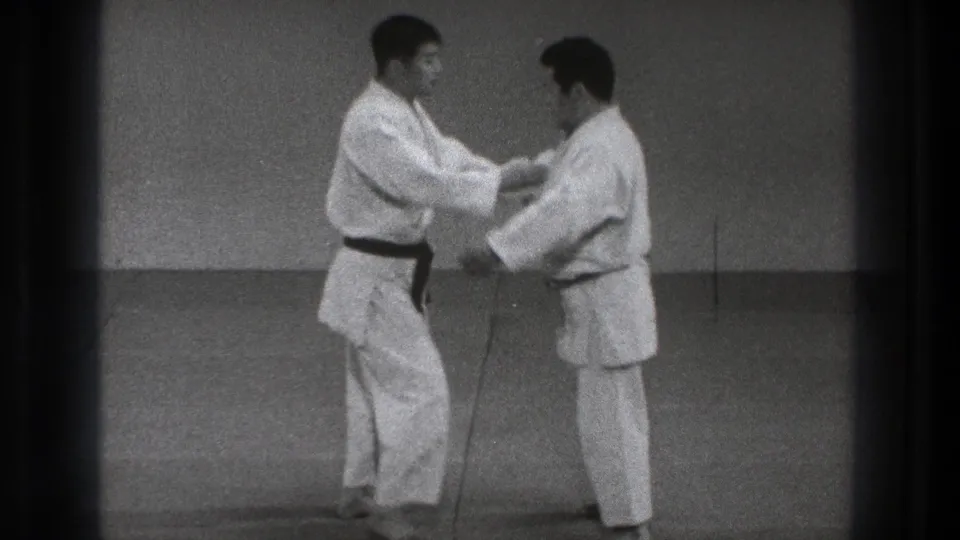Two adult men in gi make sleeve and forearm contact during a controlled martial arts technique in the 1971 Tokyo footage.