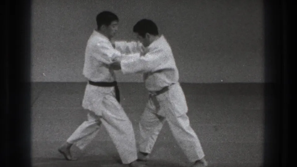 The two adult practitioners shift weight and lean into a close-range technique on the dojo mat in metadata-supported Tokyo footage.