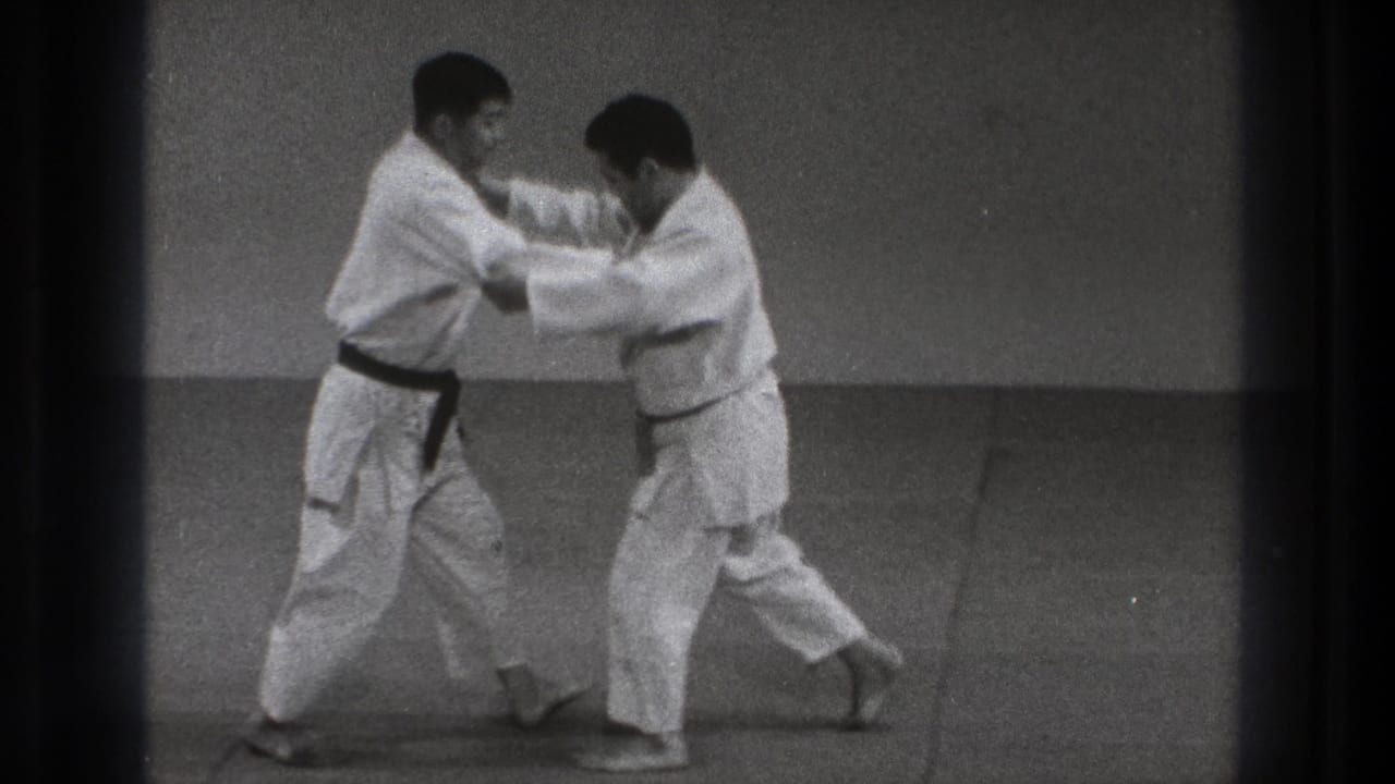 Two men in white gi grip each other while standing on an indoor mat.