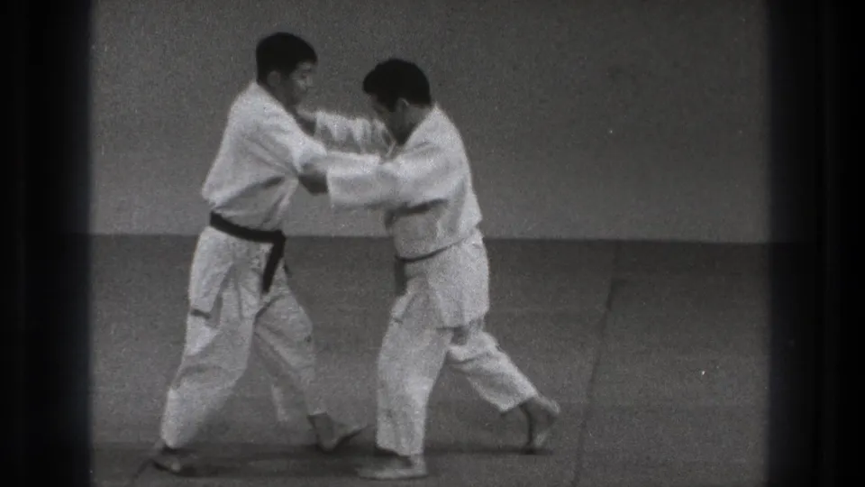 In 1971 Tokyo footage, two adult martial artists in white gi face each other on a plain indoor mat at the start of a judo throw practice.